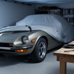 Classic Datsun Z-car parked under a garage cover with seasonal maintenance supplies like a battery tender and tire pressure gauge on a nearby workbench.